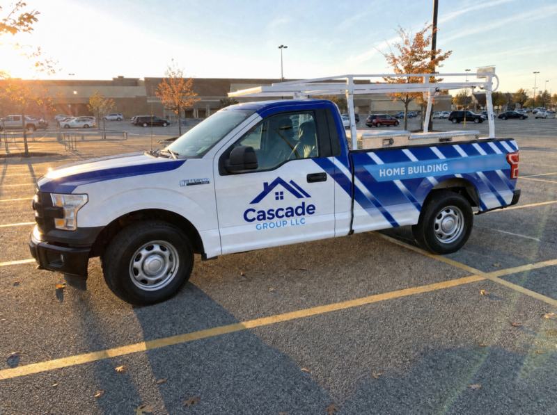 Concept mockup: white Ford F-150 pickup with a ladder rack and toolbox, driver-side profile in a store parking lot at sunset, with a navy and blue diagonal-stripe partial wrap on the bed, a blue hood stripe, a house-roof "Cascade Group LLC" logo on the door, and a "Home Building" banner.
