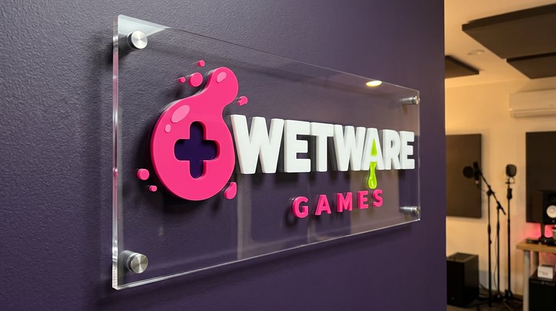 Concept mockup: clear acrylic lobby sign mounted on brushed stainless standoffs on a textured purple wall, angled view, with raised 3D lettering: a pink blob logo mark with a game-controller cross cut-out, white WETWARE with a lime-green drip from the A, and pink GAMES below, with a warmly lit recording studio with microphones and speakers to the right.